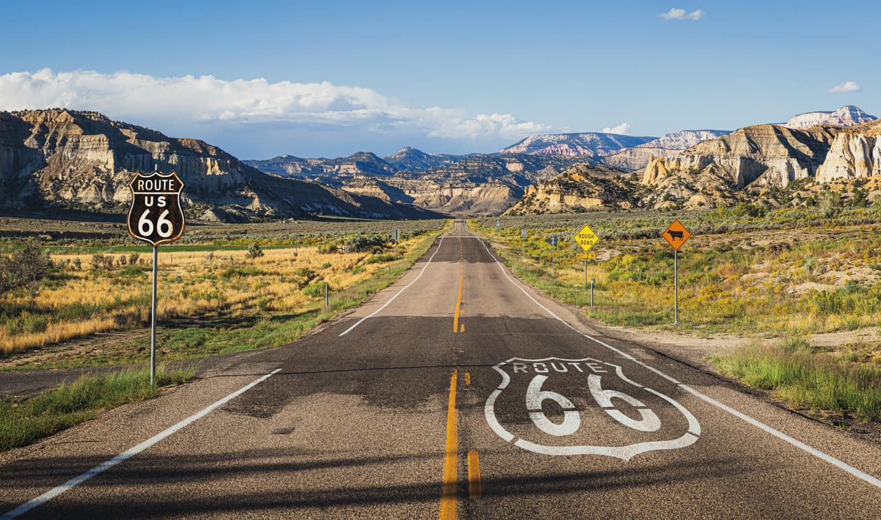 Carretera recta con el símbolo de la Ruta 66 pintado en el asfalto y montañas al fondo.