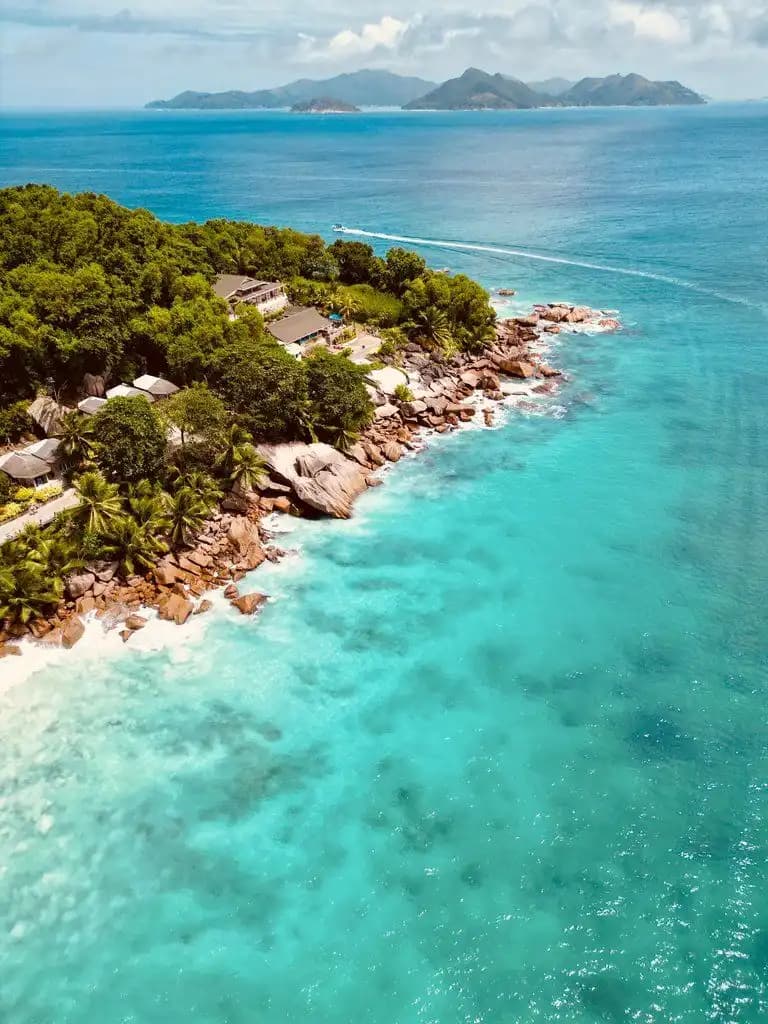 Veduta aerea di una costa rocciosa boscosa sull'acqua turchese, con una piccola barca verso colline lontane.