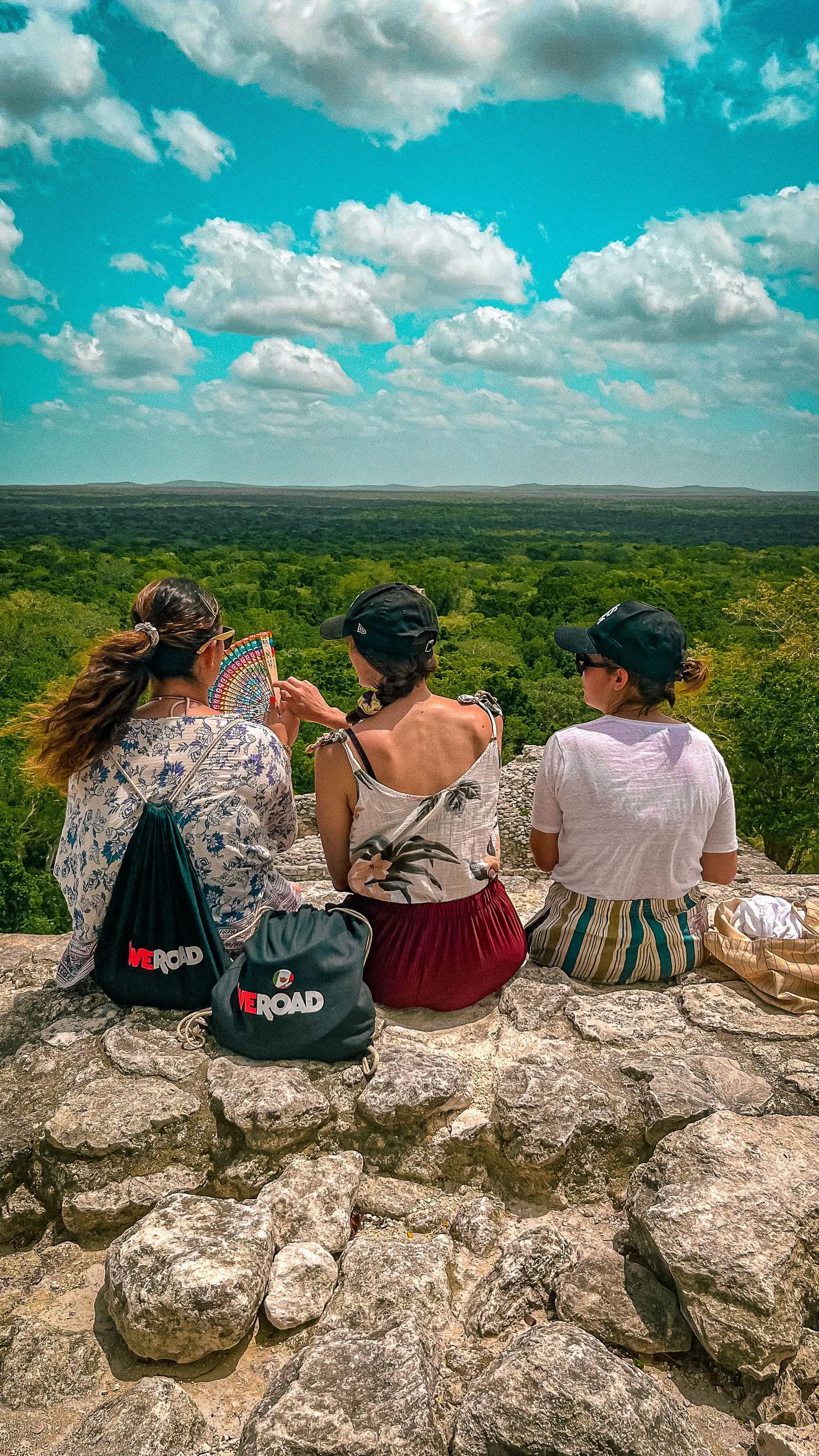 Un viaggio di gruppo WeRoad di tre persone sedute su rovine di pietra con borse WeRoad, sopra una vasta foresta.