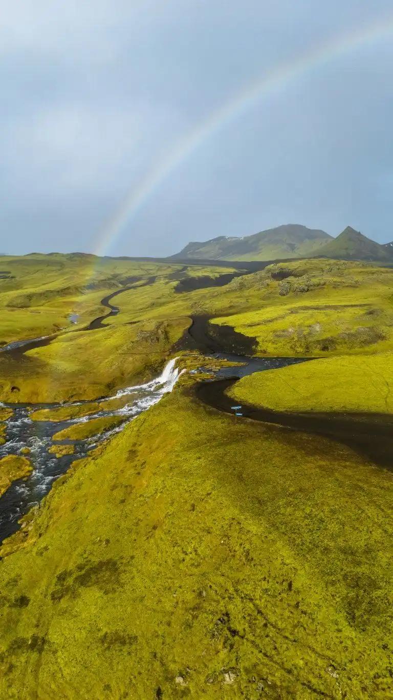 Un arcoíris sobre colinas verdes cubiertas de musgo, con un río sinuoso y una pequeña cascada.
