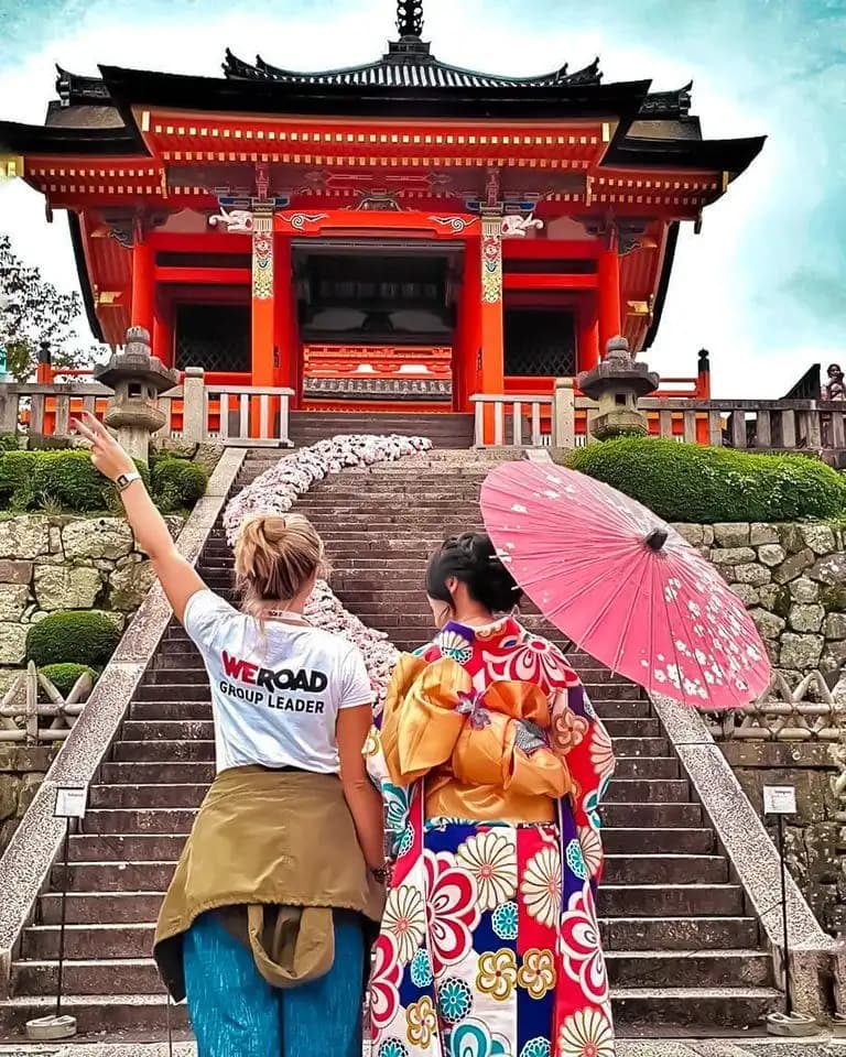 Eine WeRoad-Gruppenreise: zwei Personen vor einem verzierten roten Tempel, eine im Blumen-Kimono mit rosa Schirm.