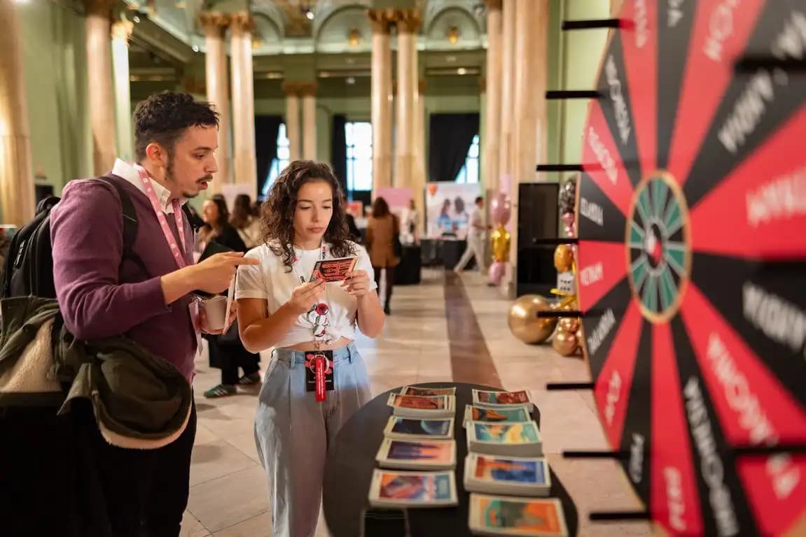 Dos personas observan una tarjeta junto a una mesa de postales de viaje y una gran rueda de la fortuna en un salón.