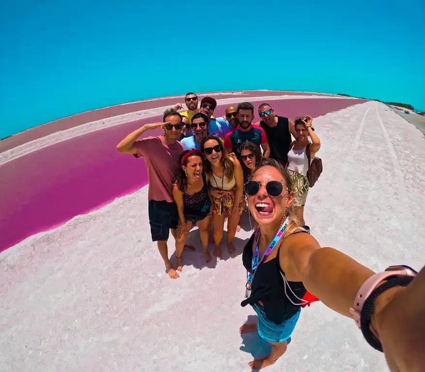 Un viaje en grupo de WeRoad haciéndose un selfie gran angular junto a un lago rosa intenso bajo un cielo azul.