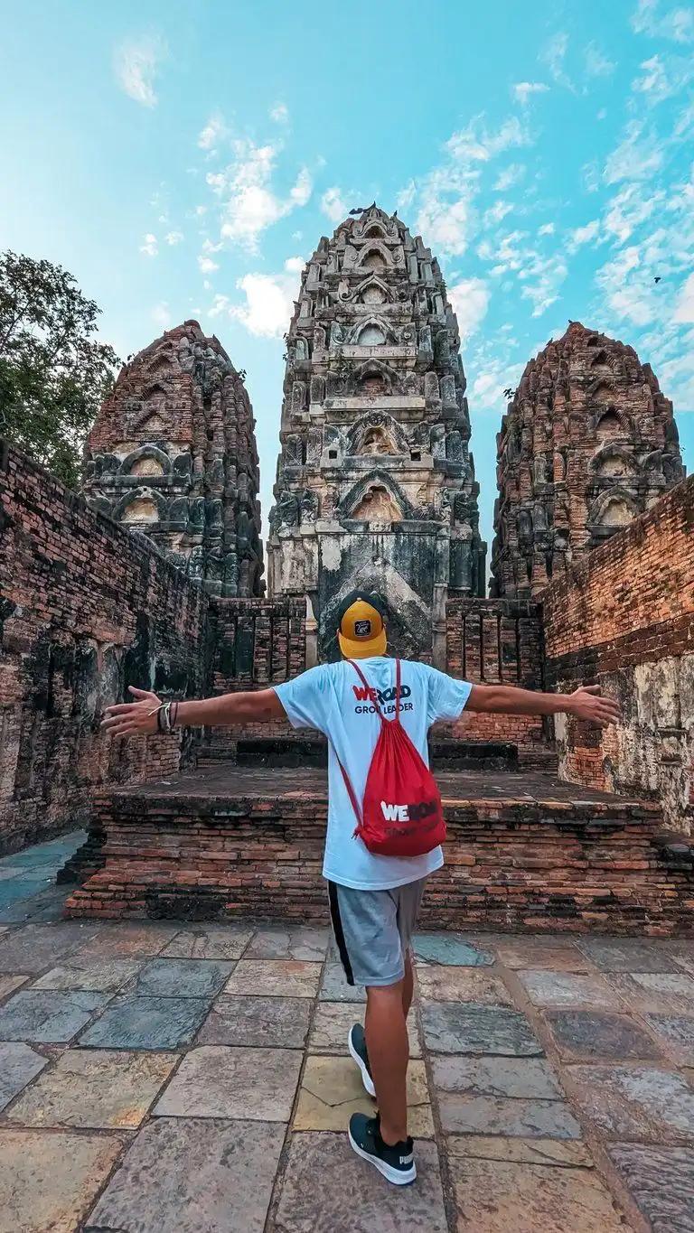 Una persona con los brazos abiertos frente a altas torres talladas de un templo, con una bolsa WeRoad roja.