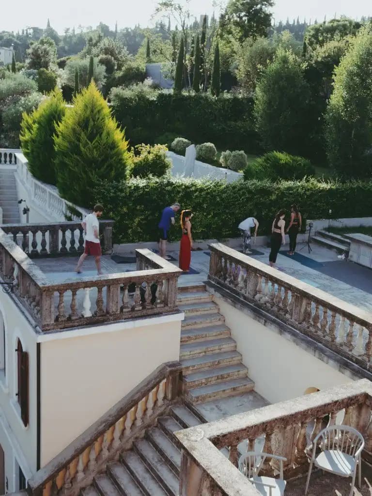 Un viaje en grupo de WeRoad haciendo yoga en la terraza de una villa, rodeado de jardines y altos cipreses.