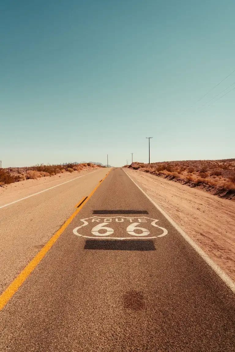 Das Route-66-Emblem auf einer leeren Wüstenstraße, die sich unter klarem Himmel zu fernen Hügeln zieht.