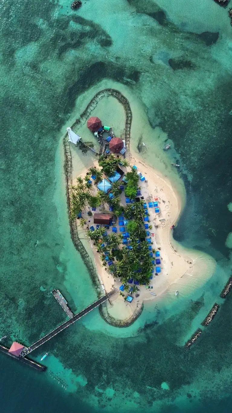 Luftaufnahme einer kleinen Sandinsel mit Palmen, Hütten und Steg, umgeben von türkisem Riffwasser.