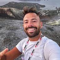 Man with a beard and mohawk wearing a WeRoad t-shirt takes a selfie with a volcanic island in the distance.