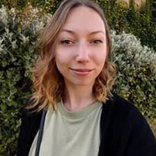 Femme aux cheveux ondulés portant une chemise verte et un cardigan foncé, devant un feuillage vert.