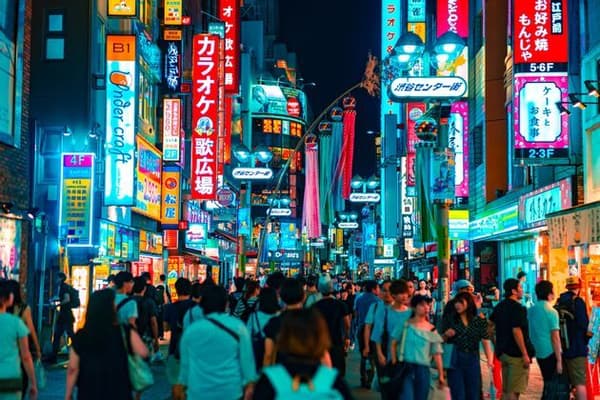 A large crowd of people walks down a narrow city street at night, illuminated by vibrant, multi-colored neon signs in Japanese.