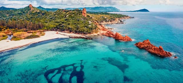 Vue aérienne d'un littoral avec une eau turquoise, une plage de sable blanc, des rochers rouges et des collines vertes sous un ciel nuageux.