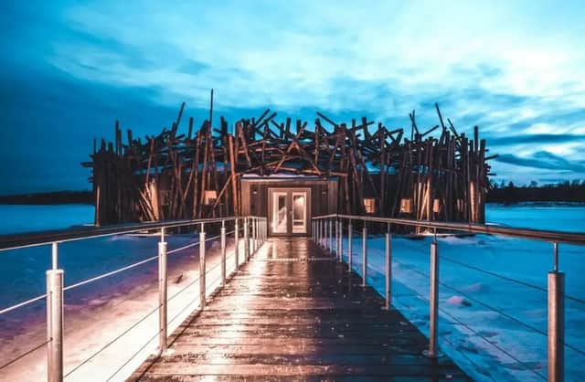 Una passerella in legno illuminata conduce a un edificio moderno con un'unica facciata a doghe, immerso in un paesaggio innevato al tramonto.