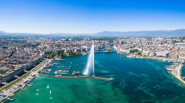 Una vista aérea de una gran fuente brotando agua desde una bahía turquesa, con una densa ciudad y montañas distantes.