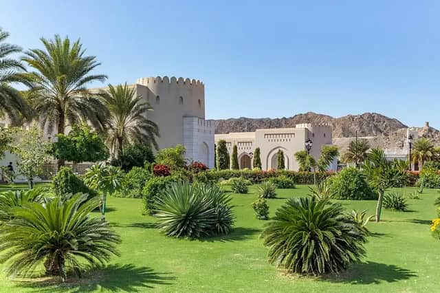 Un rigoglioso giardino verde pieno di palme e piante di fronte a un edificio tradizionale, con montagne rocciose sotto un cielo sereno.