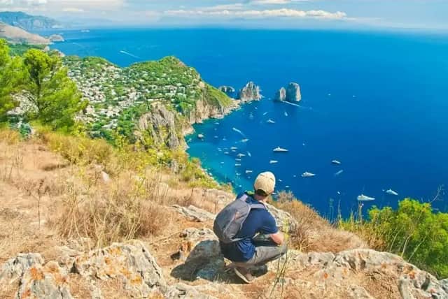 Eine Person mit Rucksack hockt auf einer felsigen Klippe und blickt auf eine Küstenstadt und ein blaues, von Booten übersätes Meer.