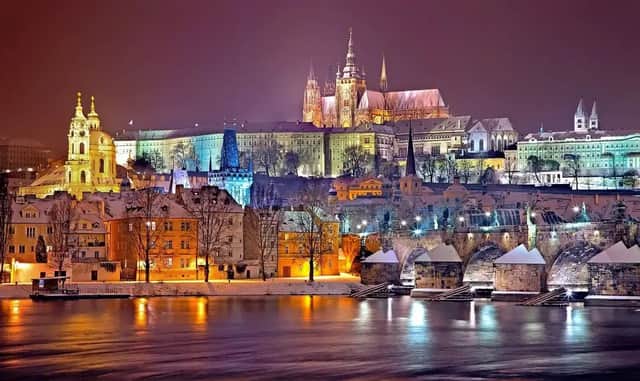 Une ville historique enneigée avec un château et un pont illuminés la nuit, les lumières se reflétant sur la rivière en contrebas.