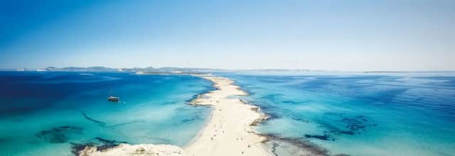 Vista aerea di una stretta lingua di sabbia che si estende in un mare turchese, con una barca a vela ancorata al largo sotto un cielo azzurro.