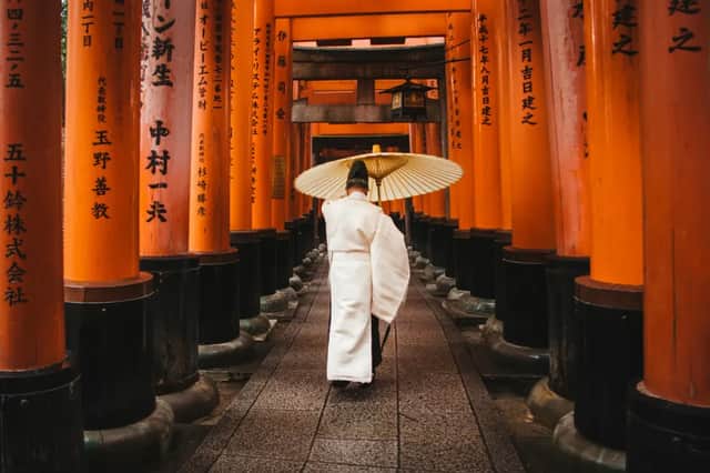 Una persona in abiti tradizionali bianchi cammina con un ombrello di carta lungo un sentiero fiancheggiato da cancelli torii arancioni.