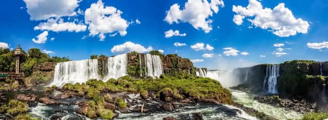 Una vista panorámica de un sistema masivo de cascadas cayendo sobre acantilados rocosos cubiertos de vegetación verde bajo un cielo azul brillante y nublado.