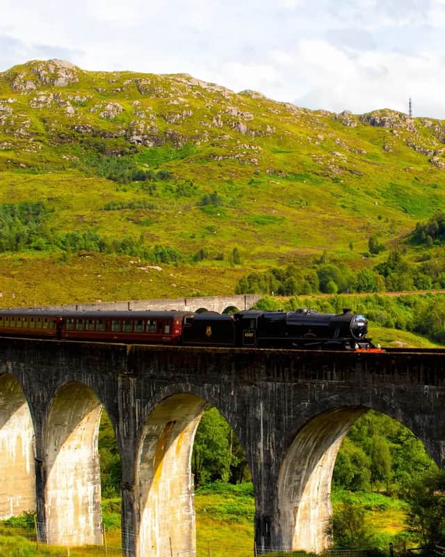 Ein schwarzer Dampfzug mit roten Waggons fährt über ein steinernes Viadukt, mit einem üppig grünen Berg im Hintergrund.