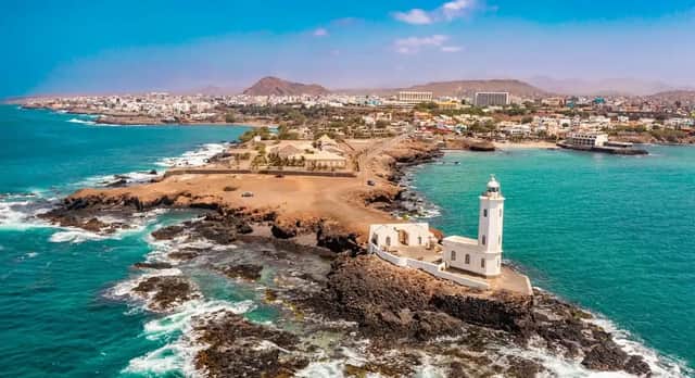 An aerial view of a white lighthouse on a rocky peninsula, with a coastal city and mountains in the background under a blue sky.