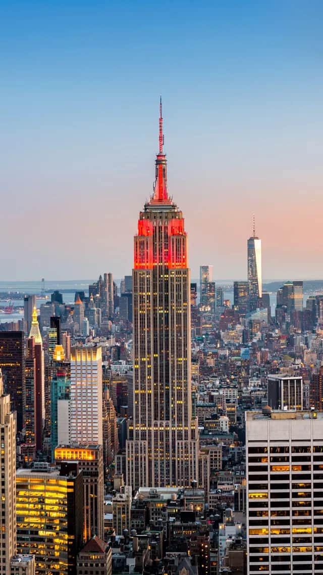 The Empire State Building illuminated with red lights at its peak, standing tall in a dense city skyline at sunset.