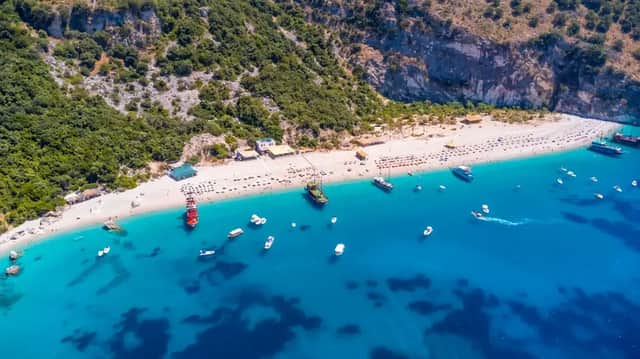 Una vista aerea di una spiaggia affollata di sabbia bianca alla base di una collina verde, con molte barche che galleggiano nel mare turchese.