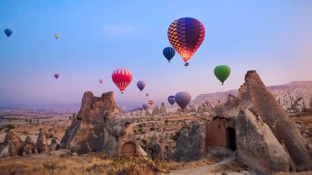 Mongolfiere colorate sorvolano un paesaggio di formazioni rocciose uniche e abitazioni rupestri durante un'alba dai colori pastello.
