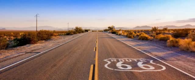 Un panneau Route 66 peint sur l'asphalte d'une autoroute vide traversant un paysage désertique avec des montagnes au loin.