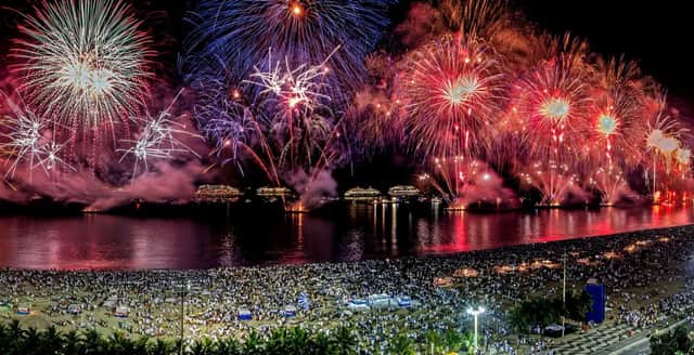 Un impresionante espectáculo de fuegos artificiales ilumina el cielo nocturno sobre una playa abarrotada de gente que mira desde la orilla.