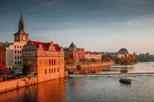 Historic buildings with red roofs and a clock tower line a river during a golden sunset, with a tour boat in the water.