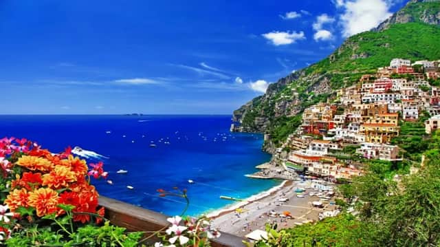 Una vista dall'alto di una colorata città costiera costruita su una scogliera verde, affacciata su una spiaggia e un mare blu profondo con barche.