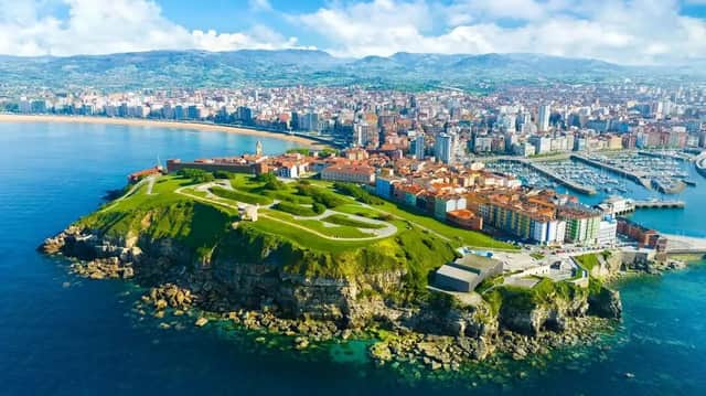Una vista aérea de una ciudad costera con una península verde, un gran puerto deportivo con barcos y una playa de arena.