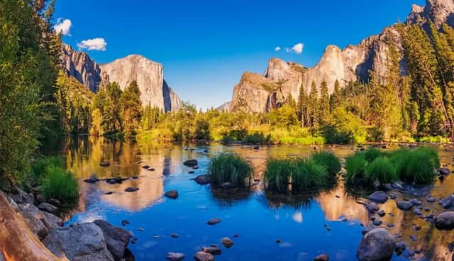 Un fiume calmo con rive rocciose riflette imponenti scogliere di granito e foreste lussureggianti sotto un cielo azzurro brillante.