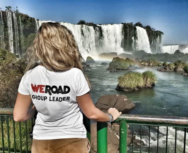 Eine blonde WeRoad Gruppenleiterin steht an einem Geländer und blickt auf einen großen, kaskadierenden Wasserfall unter klarem blauem Himmel.