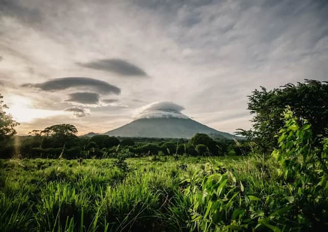 Un volcan avec un nuage lenticulaire au-dessus de son sommet, s'élevant au-dessus d'un paysage verdoyant de champs et de forêt au coucher du soleil.