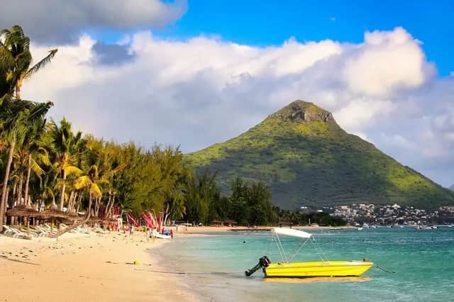 Un bateau à moteur jaune repose dans l'eau turquoise d'une plage de sable bordée de palmiers, avec une grande montagne verte en arrière-plan.