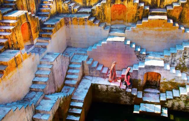 Dos mujeres con vestimenta tradicional descienden las escalinatas de piedra geométricas de un antiguo pozo escalonado.