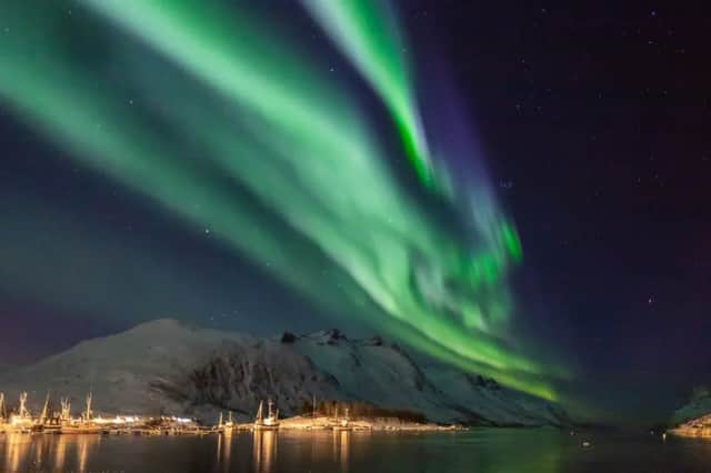 The green aurora borealis glows in the starry night sky above snow-covered mountains and a harbor with boats.
