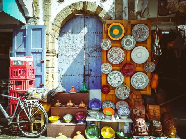 Un puesto de mercado exhibe cerámica colorida, tajines y bolsos de cuero frente a una gran puerta de madera rústica azul.