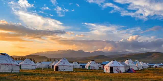 Un campamento de yurtas blancas se asienta en un campo de hierba con montañas al fondo bajo un cielo al atardecer.