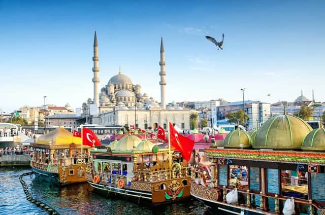Barcos ornamentados con banderas turcas atracados en el malecón de una ciudad, con una gran mezquita con minaretes al fondo.