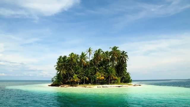 Eine kleine tropische Insel, bedeckt mit Palmen, liegt in klarem, türkisfarbenem Wasser unter einem blauen Himmel mit leichten Wolken.