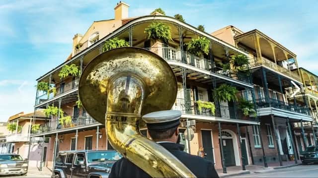 A musician from behind carries a large brass tuba on a street in front of a building with ornate, plant-filled balconies.