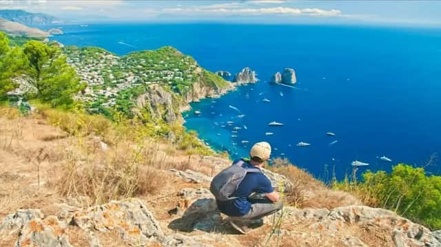 Una persona con mochila se agacha en lo alto de un acantilado rocoso, contemplando un mar azul intenso con barcos, farallones y un pueblo costero.