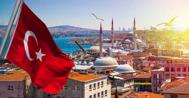Un drapeau turc flotte au-dessus d'un paysage urbain ensoleillé de dômes et de minarets, avec des mouettes survolant un plan d'eau.