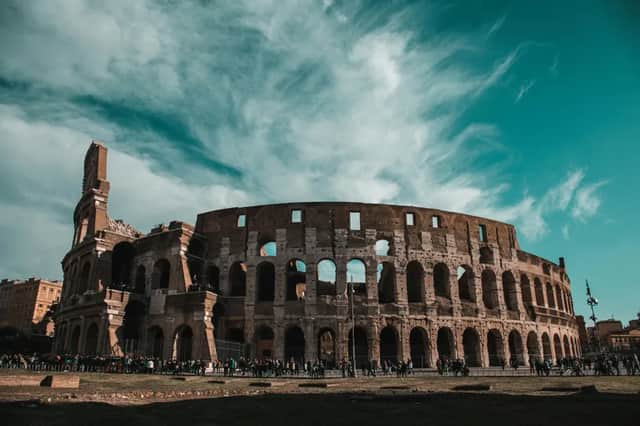 Un grand amphithéâtre antique en pierre se dresse sous un ciel sarcelle parsemé de nuages légers, avec une foule de gens rassemblée devant.