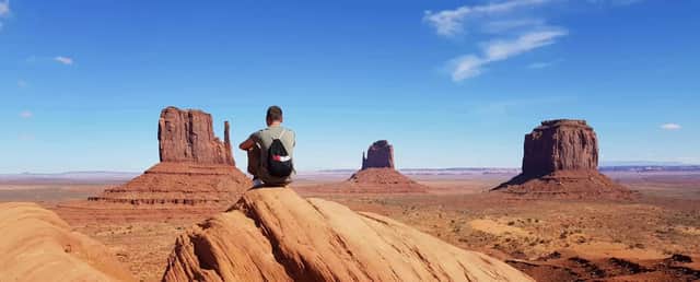 Une personne portant un sac à dos WeRoad est assise sur un rocher rouge, dominant une vallée désertique avec de grandes buttes sous un ciel bleu.