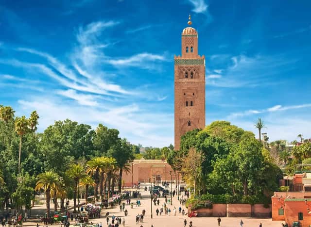Un minaret élancé couleur sable domine une place de ville animée, remplie de monde et de palmiers, sous un ciel bleu.