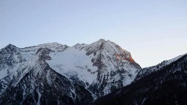 Un sommet enneigé est illuminé par la lumière orange du lever du soleil sous un ciel clair, avec des pentes boisées sombres en contrebas.